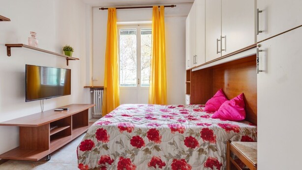 A bedroom with a bed, a TV, a window with curtains, and a radiator.