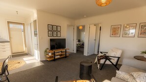Un salon comprenant un téléviseur, un canapé et des fauteuils, un coin repas avec une table et des chaises, et une cuisine équipée de placards et d’un évier.