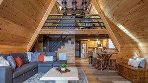 Een ruime woonruimte in loft-stijl met een hoge plafond, houten balken, een grijze hoekbank, een salontafel en een keuken op de achtergrond.