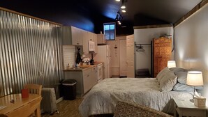 A compact hotel room with a bed, bedside table, chair, desk, kitchenette, and a small bathroom. - Downtown Fairview BunkHouse (Fairview)