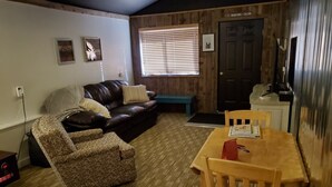 A cozy living room with a brown leather sofa, patterned armchair, and wooden dining table. A wooden door labeled "Mountains Calling" is visible in the background. - Downtown Fairview BunkHouse (Fairview)