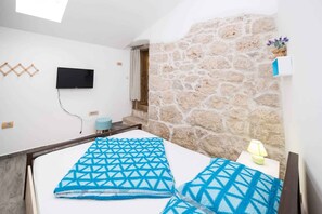 A bedroom with a stone wall, a TV mounted on the wall, a bed with blue patterned bedding, a small bedside table with a lamp, and a window with curtains. - Holiday home in outdoor pool (Tribalj)