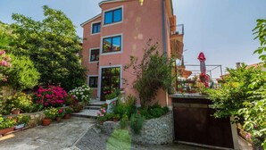 Un immeuble à plusieurs étages, doté d’une façade rose, d’un balcon à l’étage supérieur et d’un petit jardin agrémenté de plantes et de fleurs en pot.