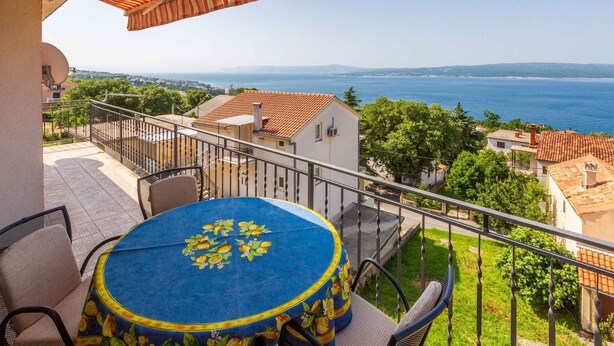 Un balcon avec une table ronde et des chaises, offrant une vue sur la côte, avec des maisons et un ciel dégagé.