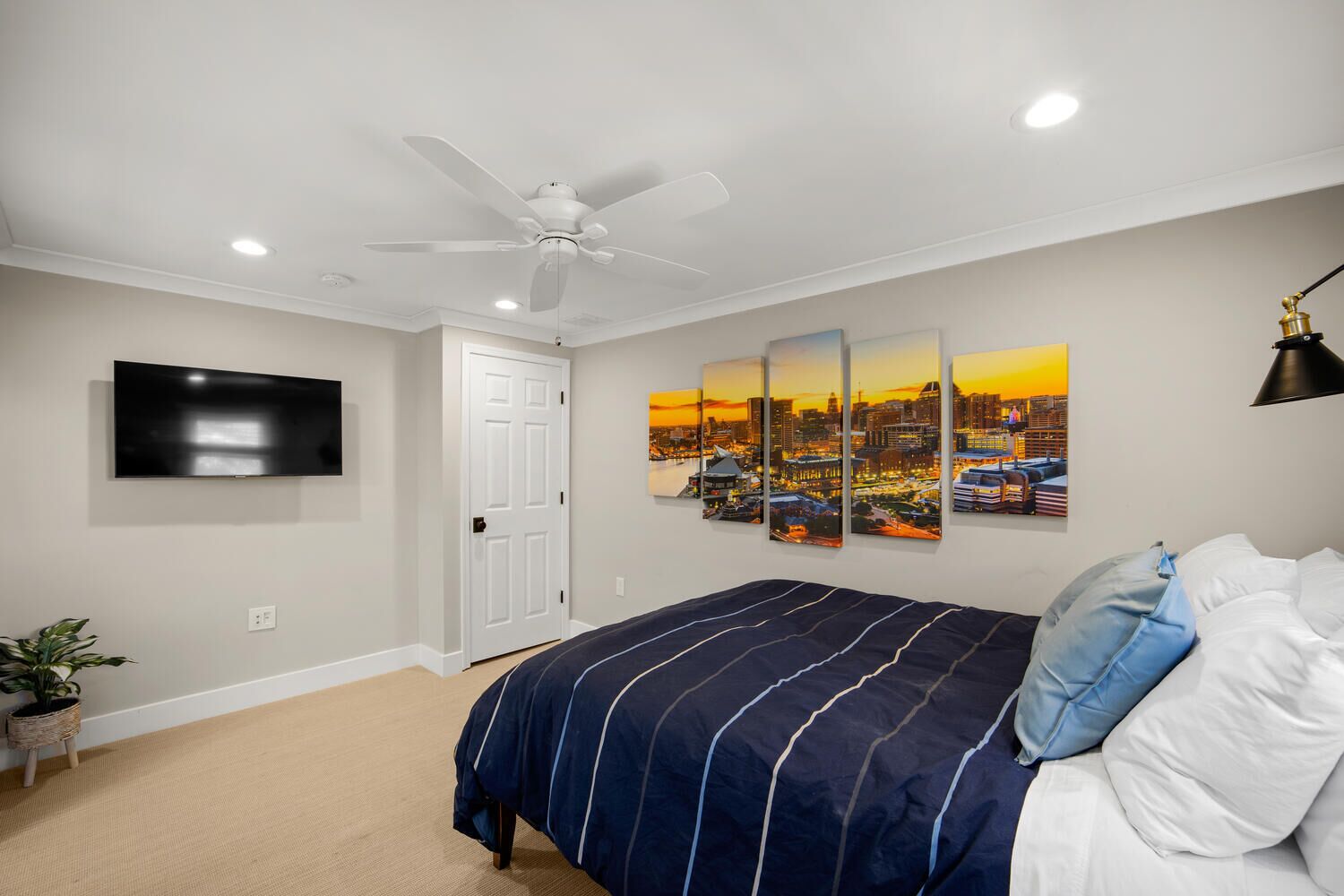 A bedroom with a bed, a ceiling fan, a TV, a door, and a cityscape painting.