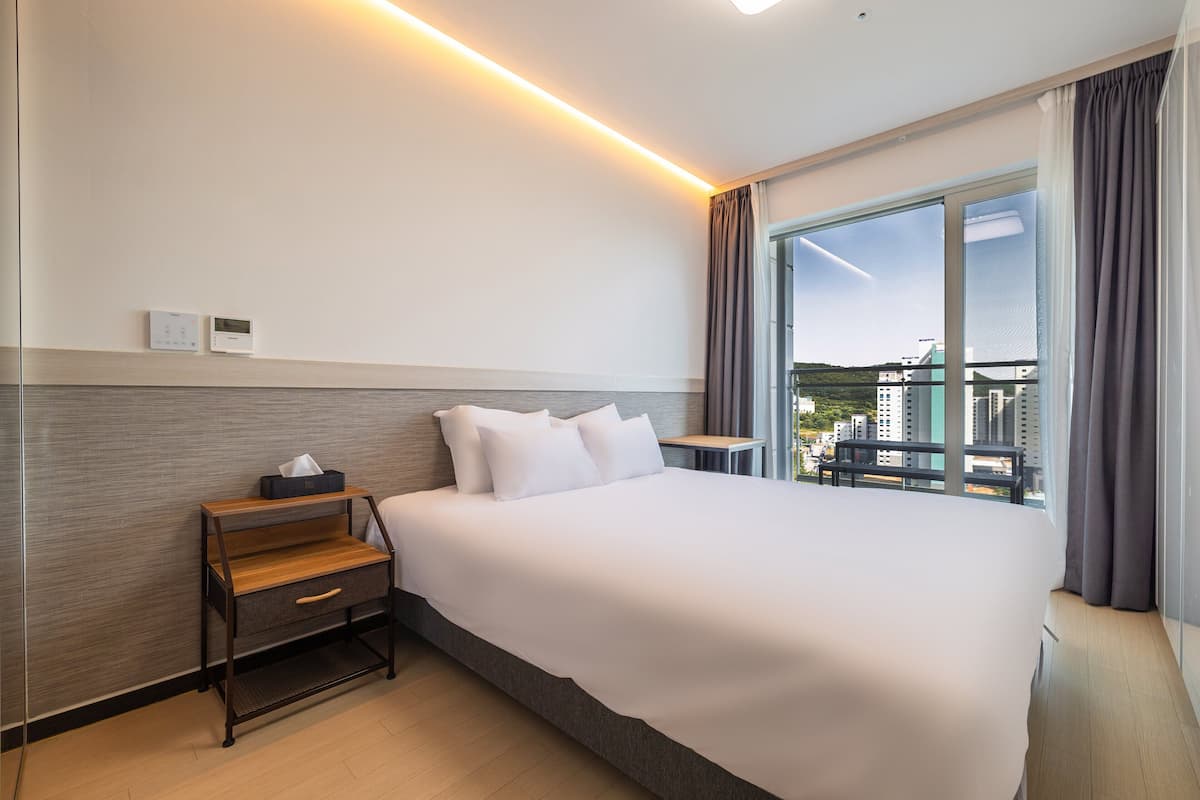 A hotel room with a large bed, a bedside table with a lamp, a thermostat, and a view of buildings through a large window.