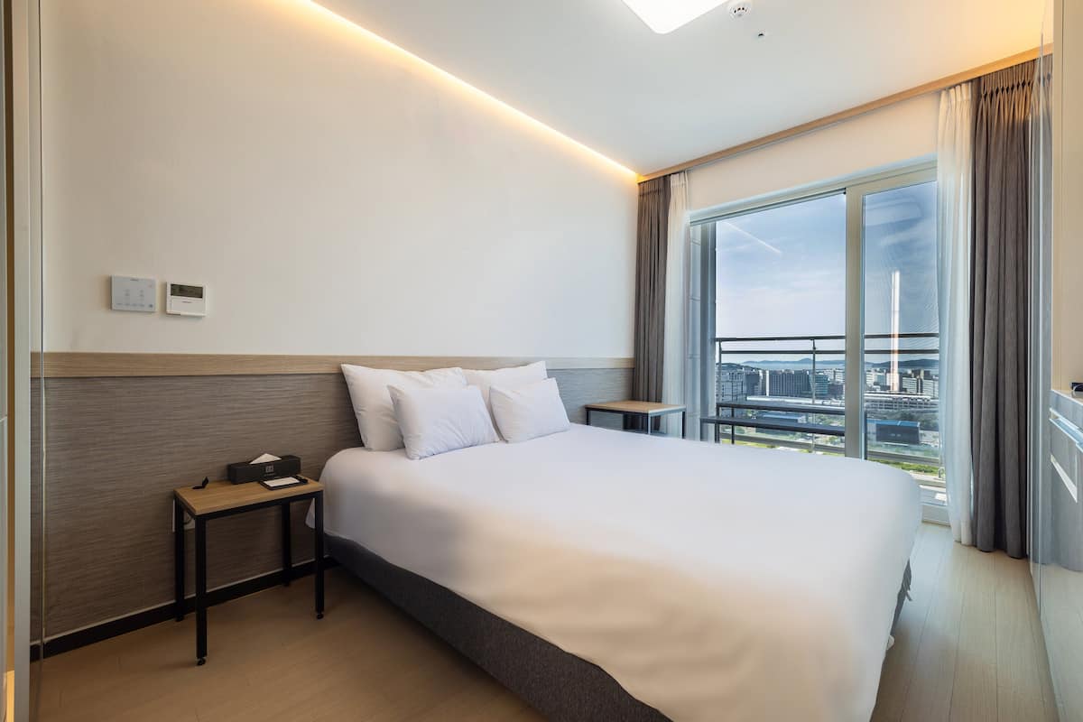 A hotel room with a large bed, a bedside table, a nightstand, a window with a city view, and a thermostat on the wall.