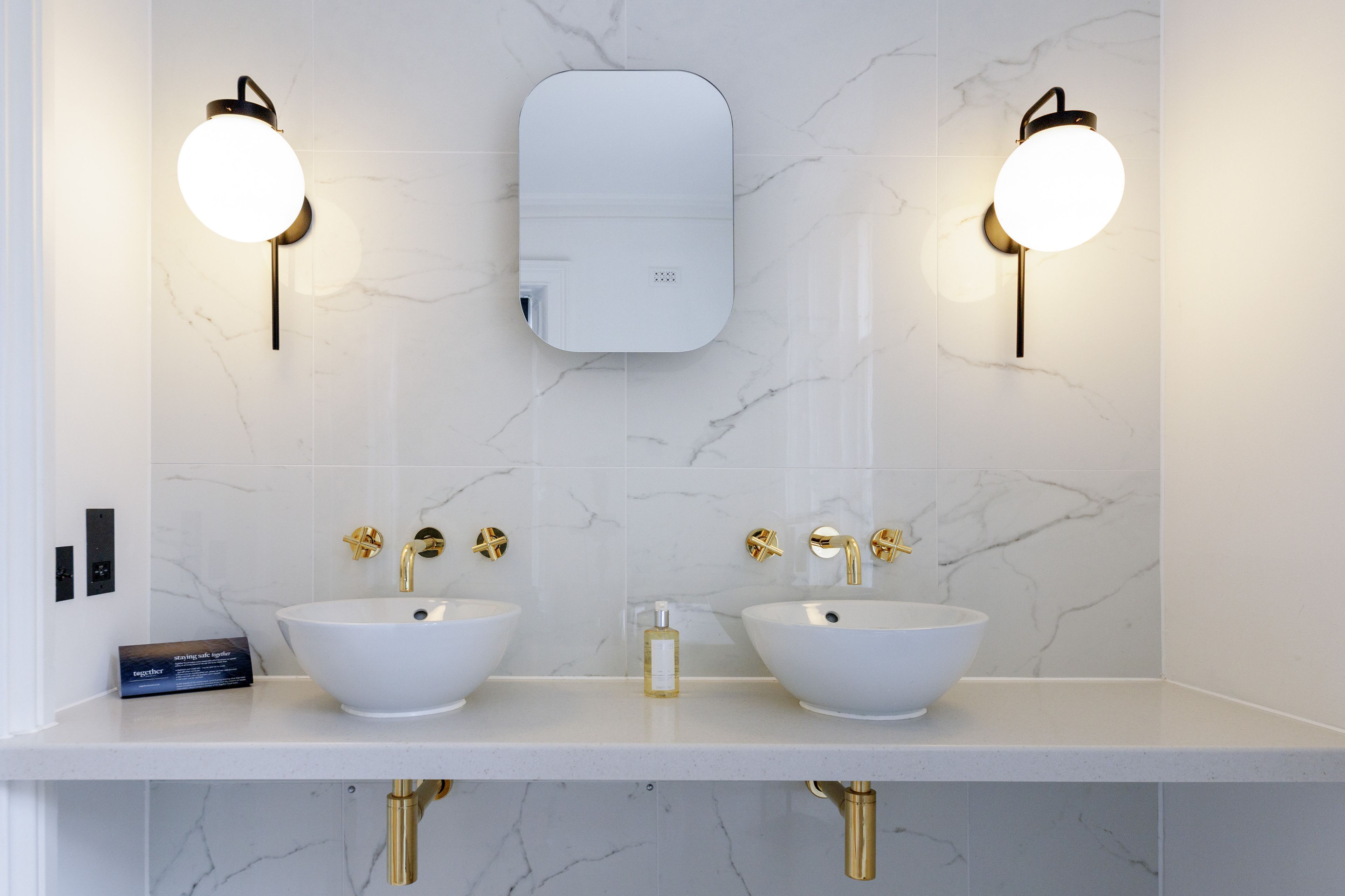 Un baño con dos lavamanos blancos, grifos dorados y apliques de pared redondos.