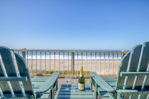 Une terrasse avec deux fauteuils Adirondack verts, une bouteille de champagne et deux verres posés sur une table, donnant sur une plage bordée d’une rambarde en bois.