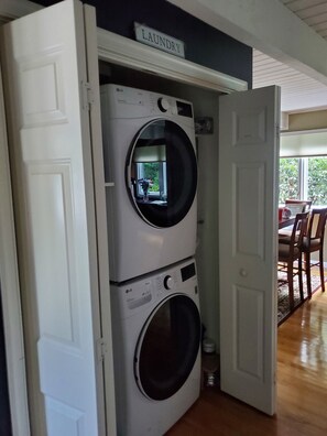 Uma lavanderia com máquinas de lavar e secar empilhadas, uma placa indicando o local e vista para uma sala de jantar com cadeiras e uma mesa.