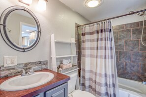 A bathroom with a round mirror, a sink, a shower with a striped curtain, and a shelf with toiletries. - Nehalem House (Nehalem)
