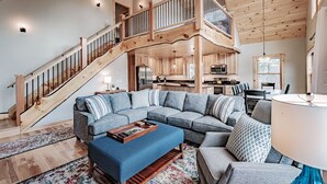 A spacious living area with a large sectional sofa, a wooden staircase, and a kitchen in the background. - Chalet Tourmaline - Charming Newly Built Chalet (Bethel)