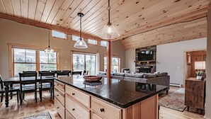 A spacious kitchen with wooden cabinets, a granite countertop, and a large island. There is a dining area with a table and chairs, and a living area with a sofa and a TV. - Chalet Tourmaline - Charming Newly Built Chalet (Bethel)