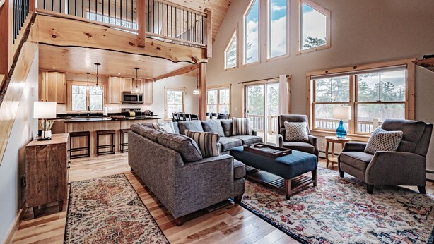 A spacious living area with a high ceiling, wooden beams, a built-in kitchen, and large windows. - Chalet Tourmaline - Charming Newly Built Chalet (Bethel)