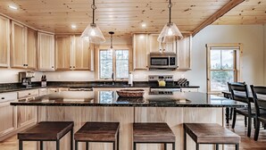 A modern kitchen with wooden cabinets, a central island with stools, stainless steel appliances, and pendant lighting. - Chalet Tourmaline - Charming Newly Built Chalet (Bethel)