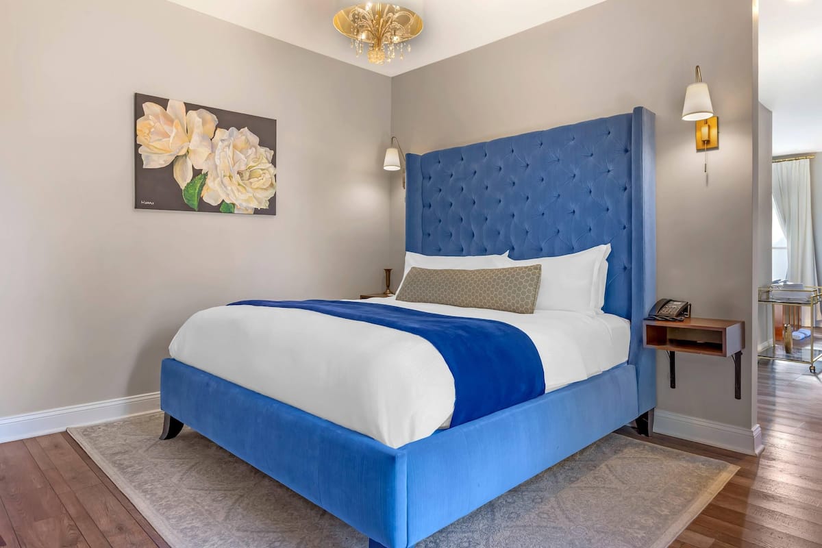 A bedroom with a large bed, a painting on the wall, a chandelier, and a nightstand with a lamp.