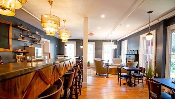 A bar area with wooden bar stools, pendant lighting, and a counter with a glass display. There are tables and chairs in the background, and the room has a wooden floor. - The Colonial Inn, an Ascend Collection Hotel (Hillsborough)