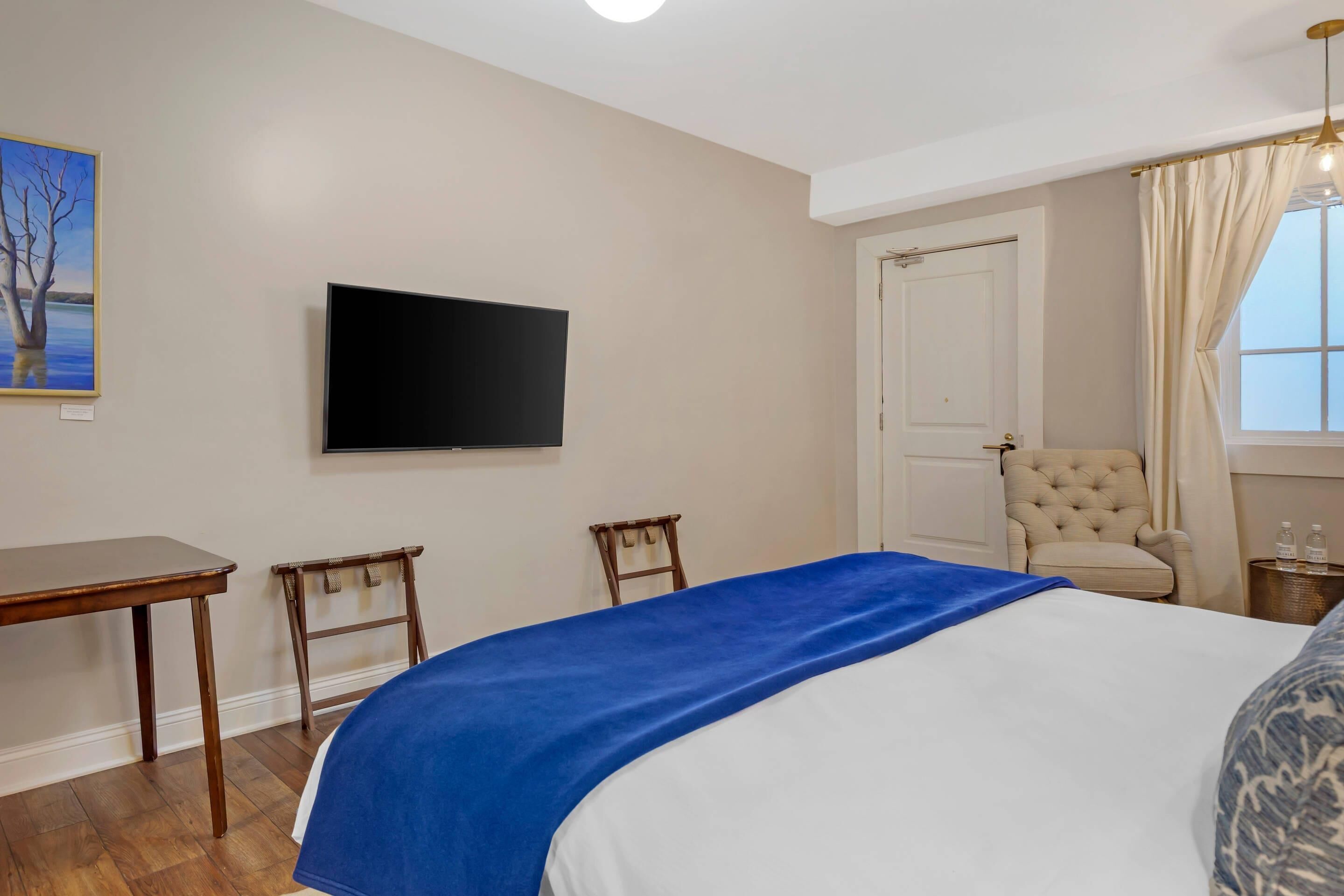 A hotel room with a bed, a flat-screen TV, a desk, a chair, a window with curtains, and a door.