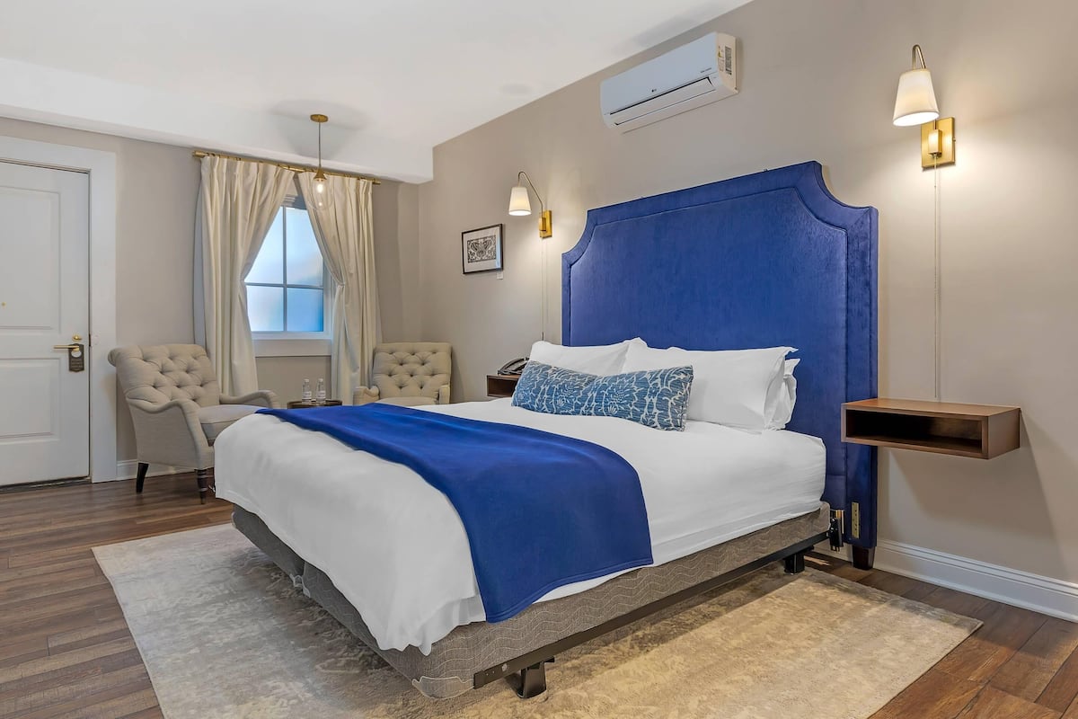 A bedroom with a bed, a chair, a nightstand, a wall-mounted shelf, and an air conditioning unit.