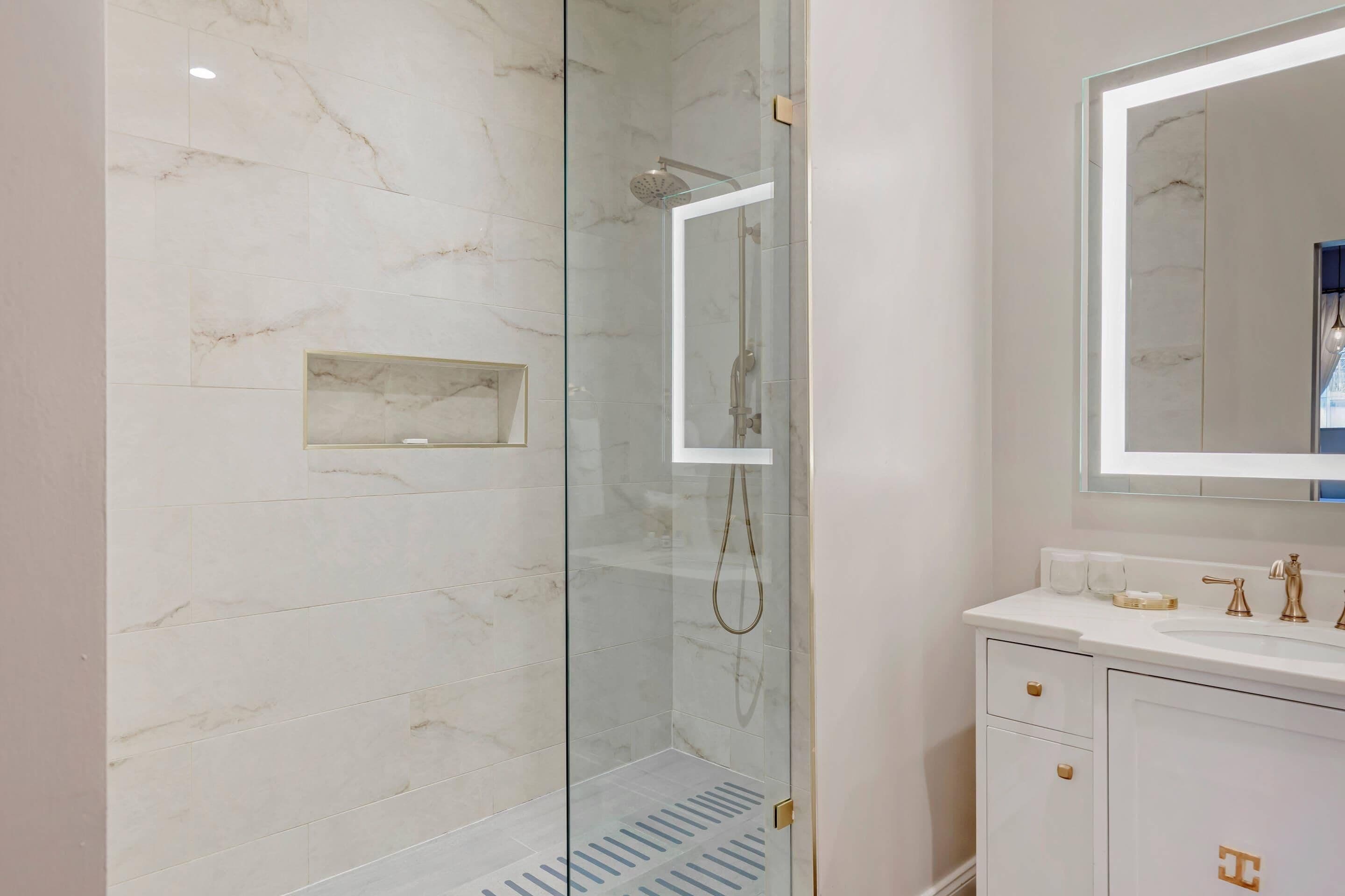 A modern bathroom with a glass-enclosed shower, a marble-tiled wall, a white vanity with gold fixtures, and a large mirror.