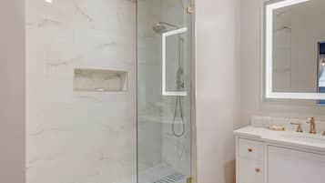 A modern bathroom with a glass-enclosed shower, a marble-tiled wall, a white vanity with gold fixtures, and a large mirror.