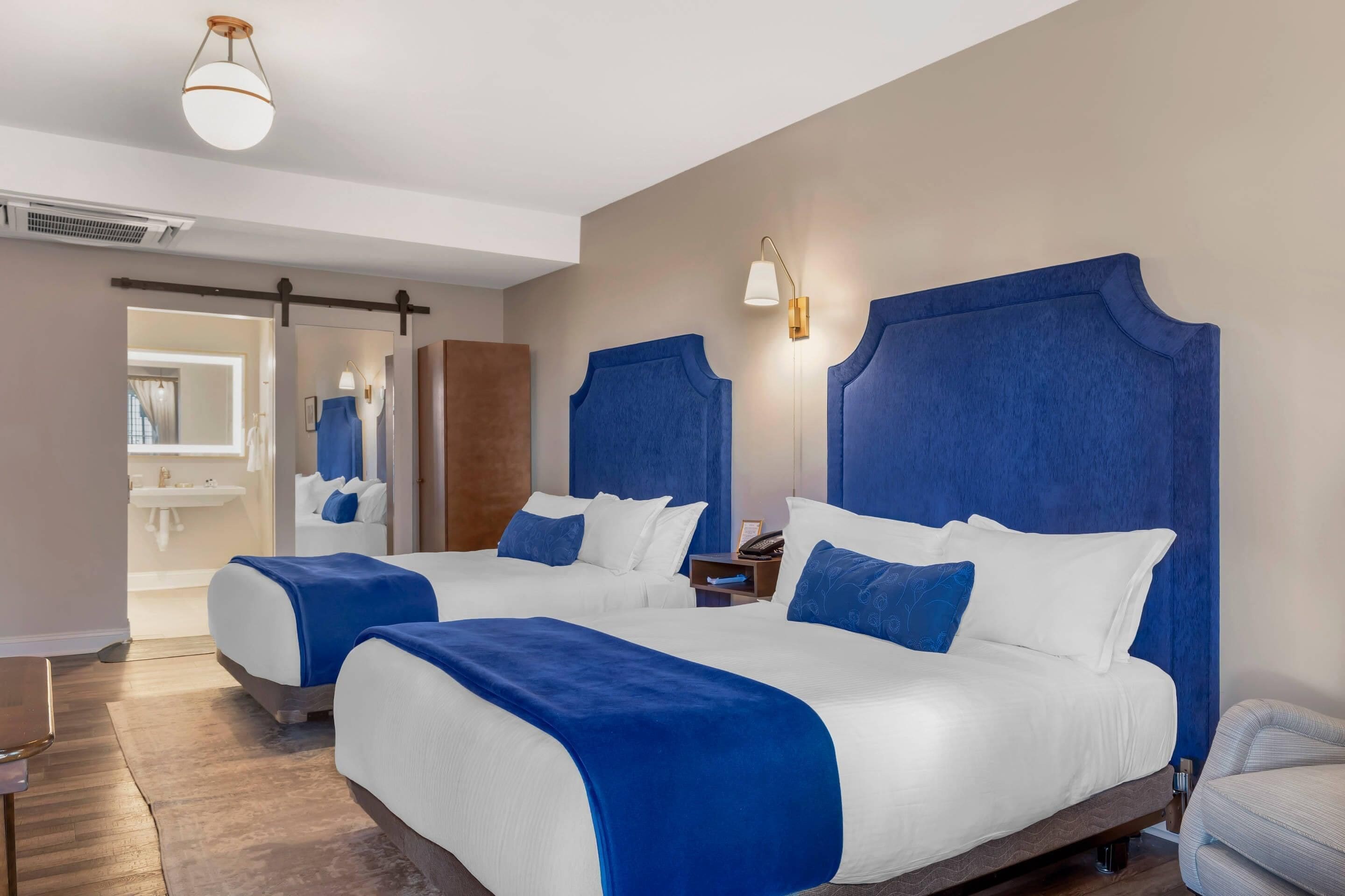 A hotel room with two beds, a bedside table, a chair, a mirror, and a bathroom visible through an open door.