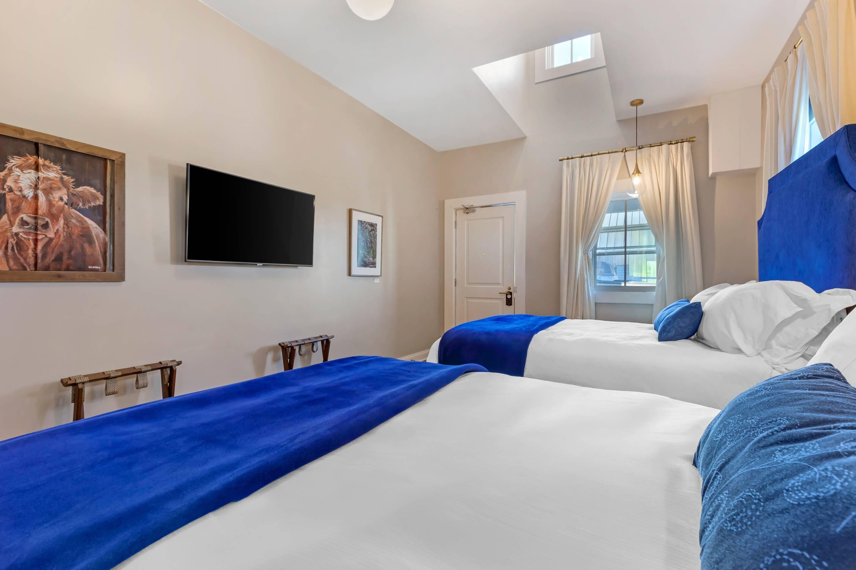 A hotel room with two beds, a TV, a painting, and a window with curtains.
