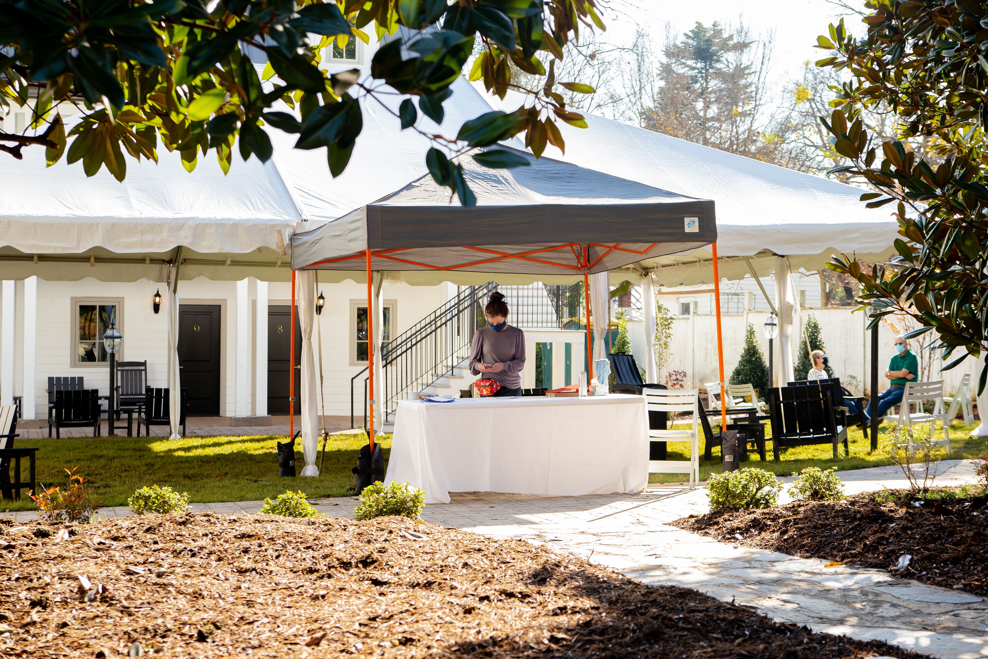 A covered outdoor area with a table and chairs, a small garden, and a building in the background.