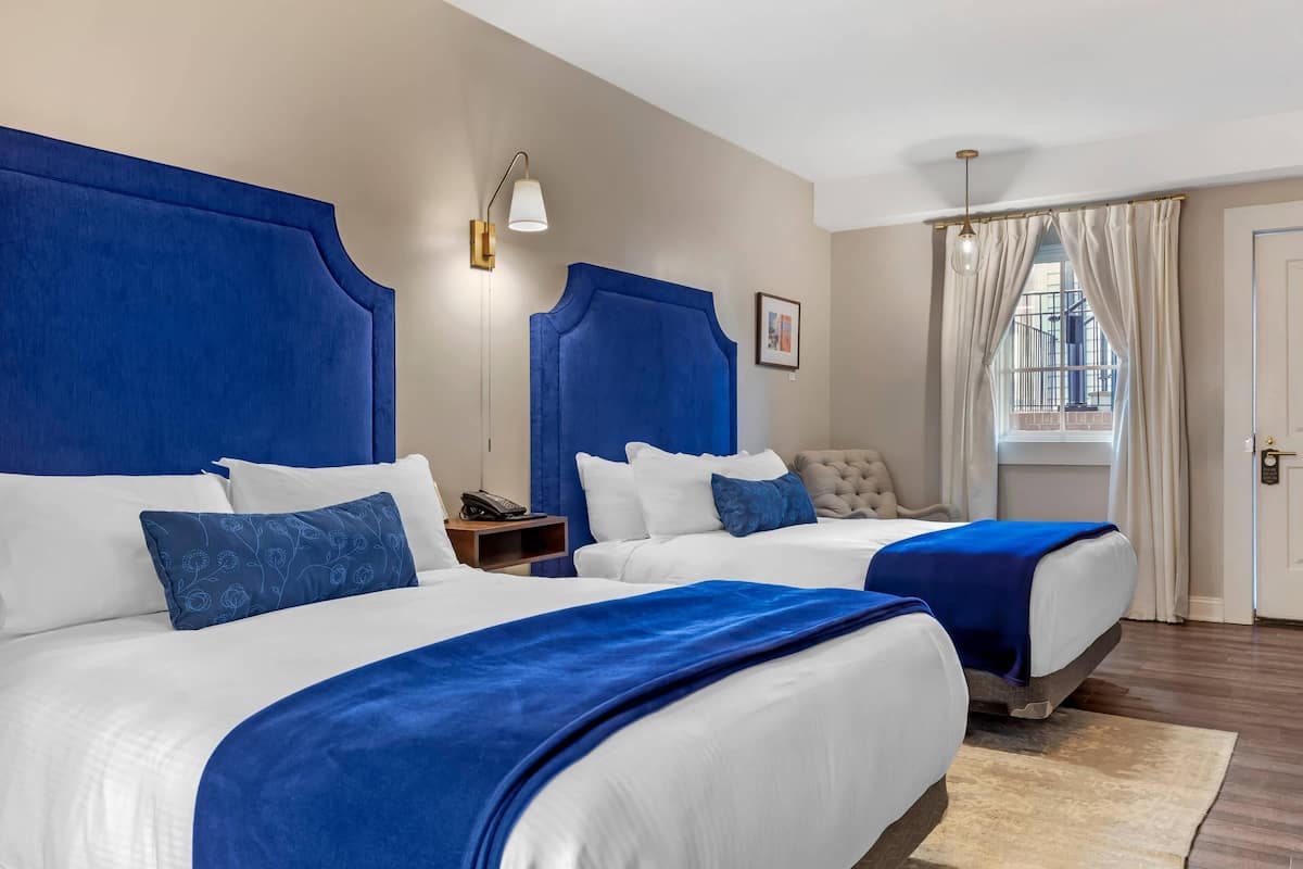A hotel room with two beds, a nightstand, a lamp, a chair, a window with curtains, and a door.
