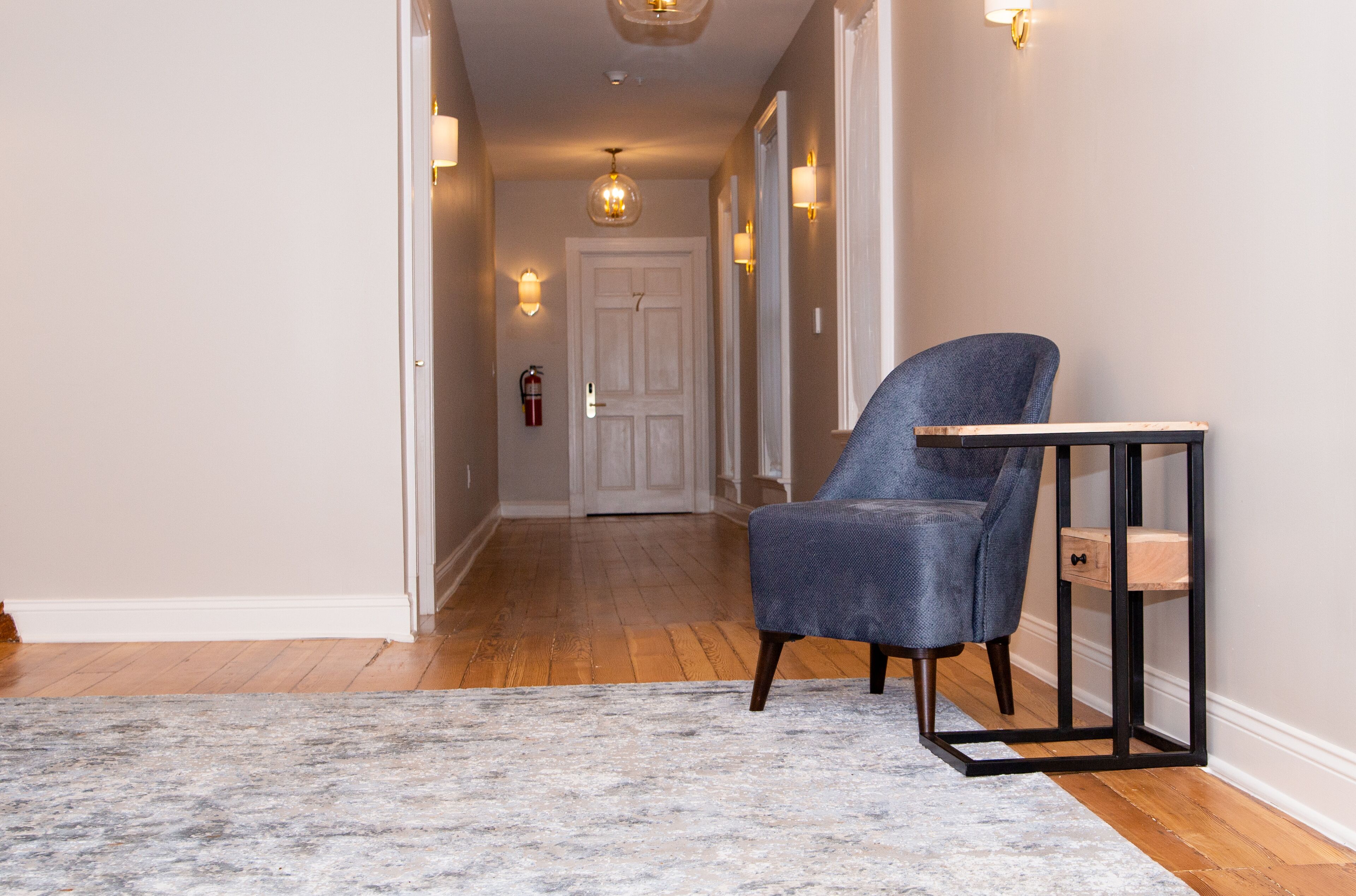 Un couloir avec un fauteuil bleu, une petite table et un tapis à motifs.