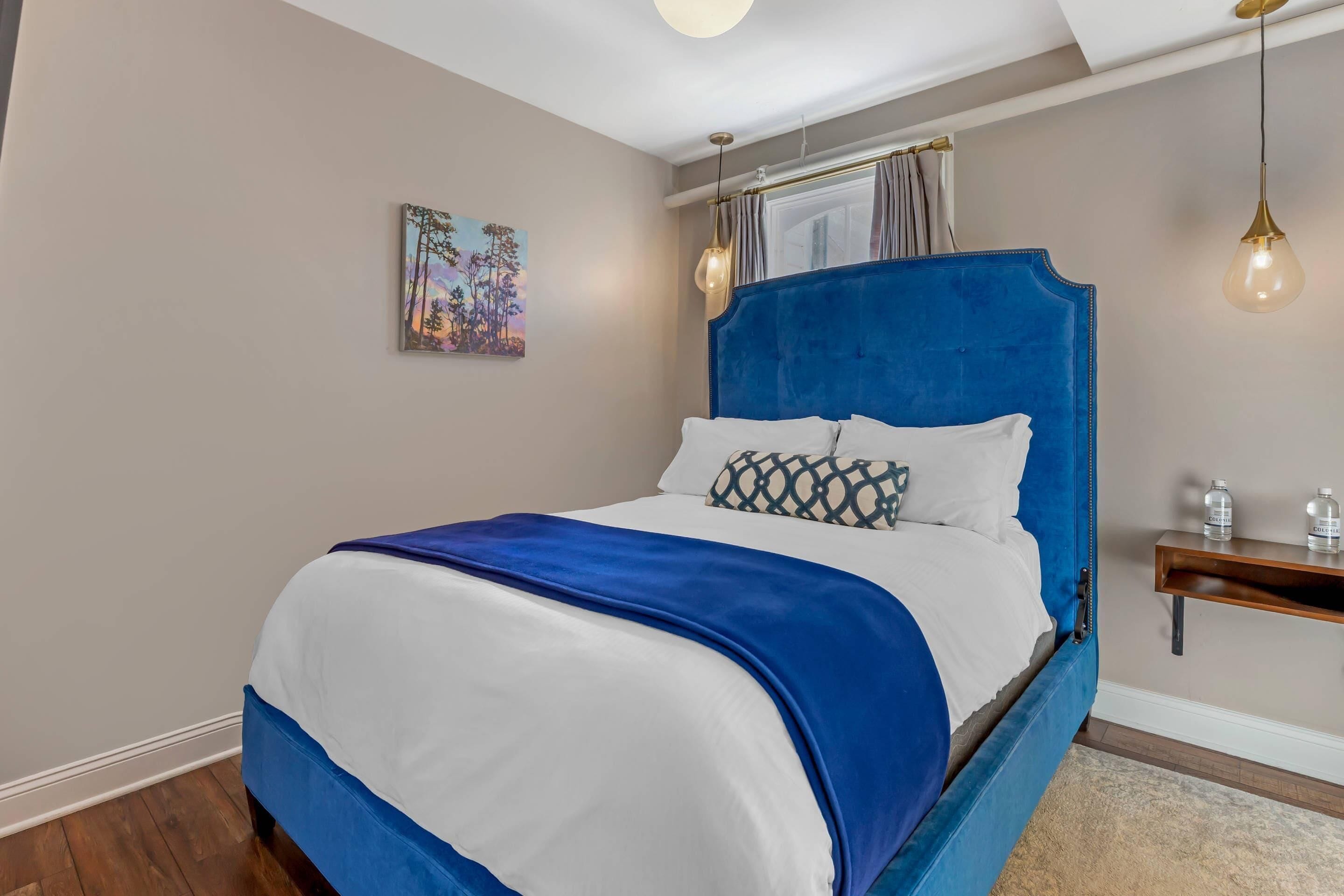 A bed with a blue headboard, white bedding, and a blue throw blanket. There is a painting on the wall, a bedside table with two bottles, and a hanging light fixture.