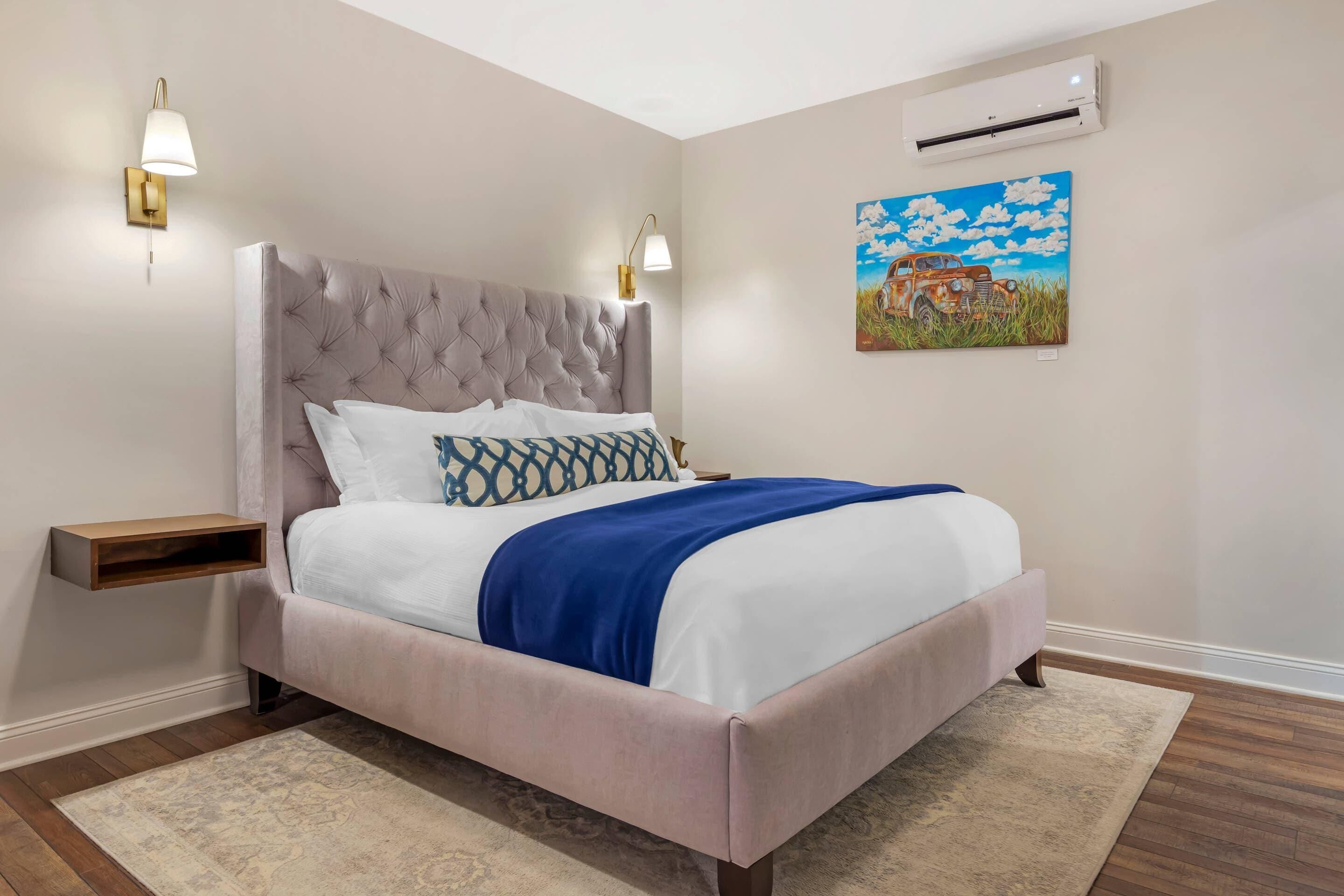 A bedroom with a tufted headboard, a bed with white and blue bedding, a blue throw blanket, a nightstand with a lamp, and an air conditioning unit on the wall.