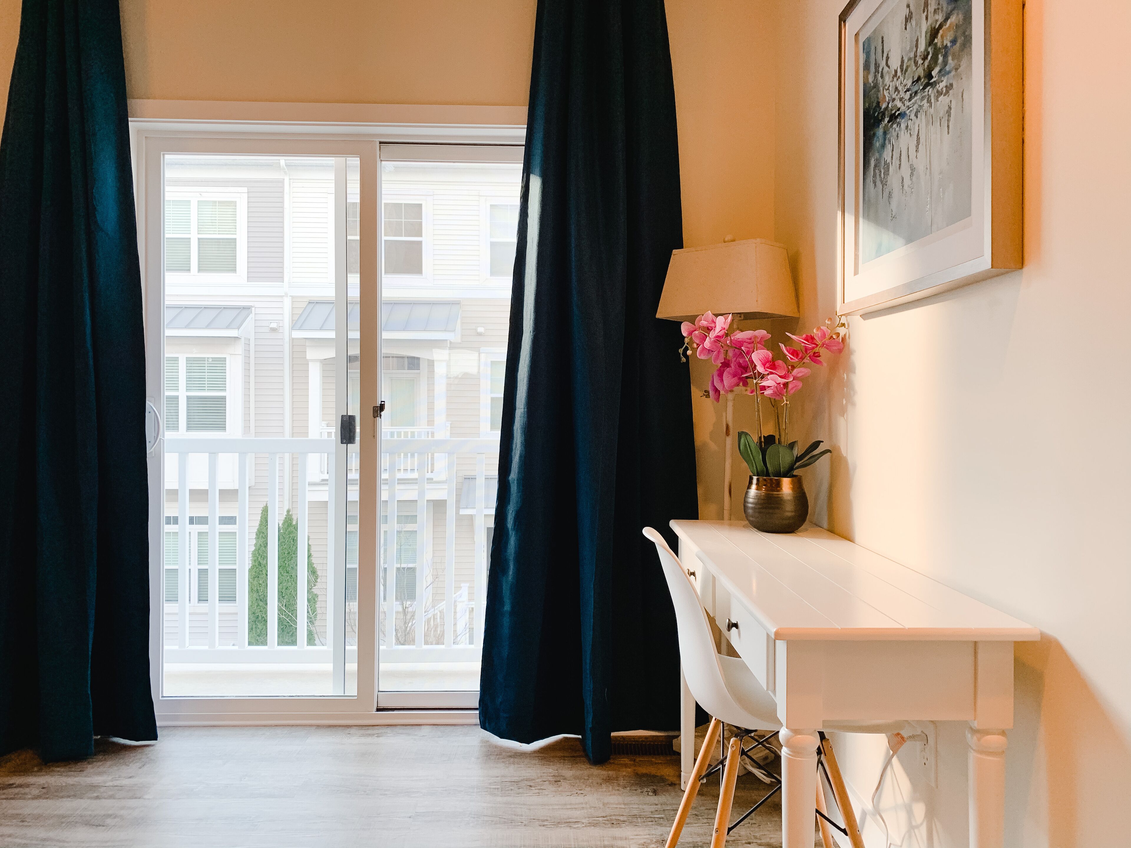 A room with a white desk, a lamp, a vase with pink flowers, and a view of a building through a glass door.