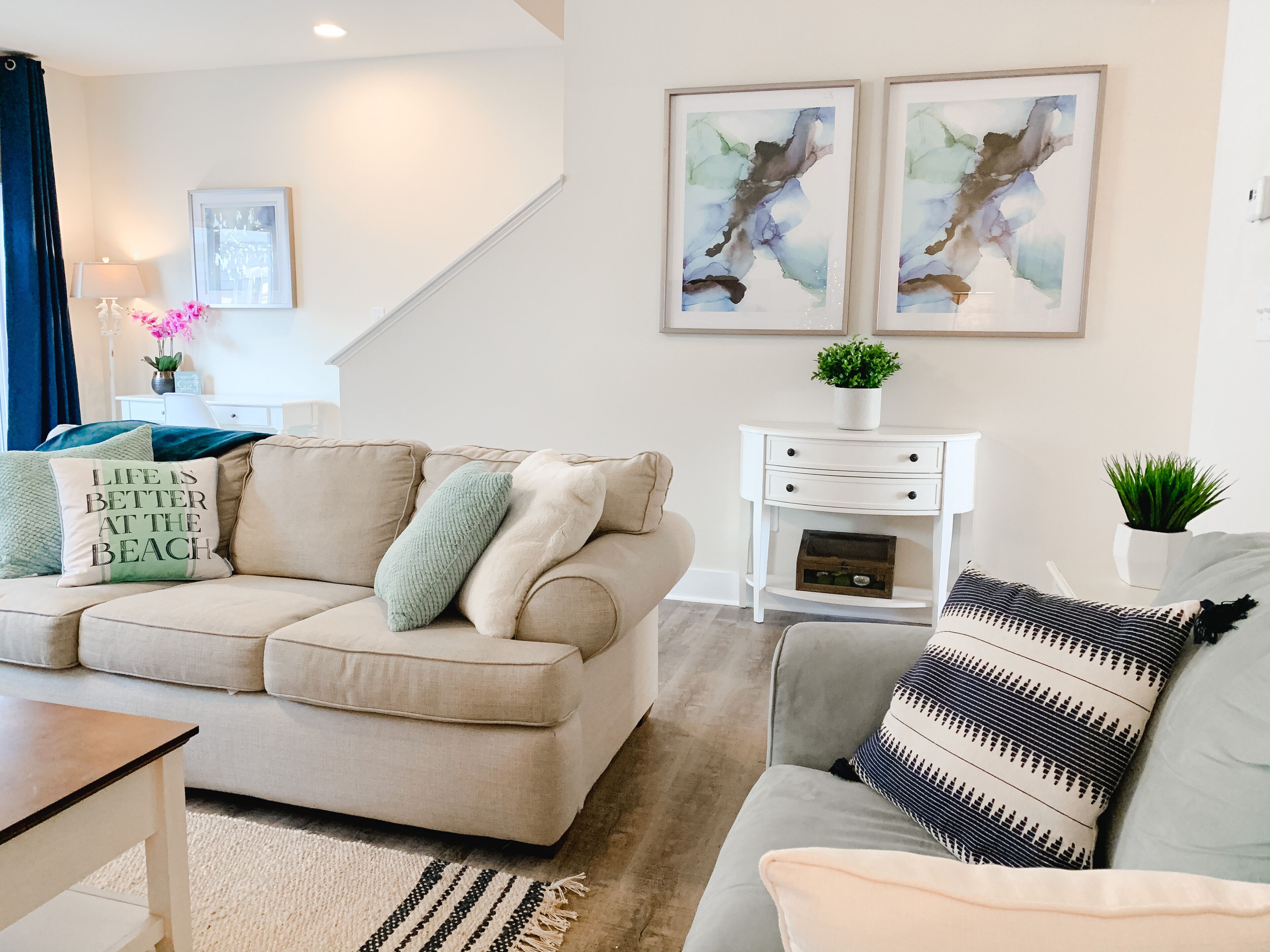 A living room with a beige sofa, a grey sofa, a white side table, and a coffee table. There are two framed abstract paintings on the wall.