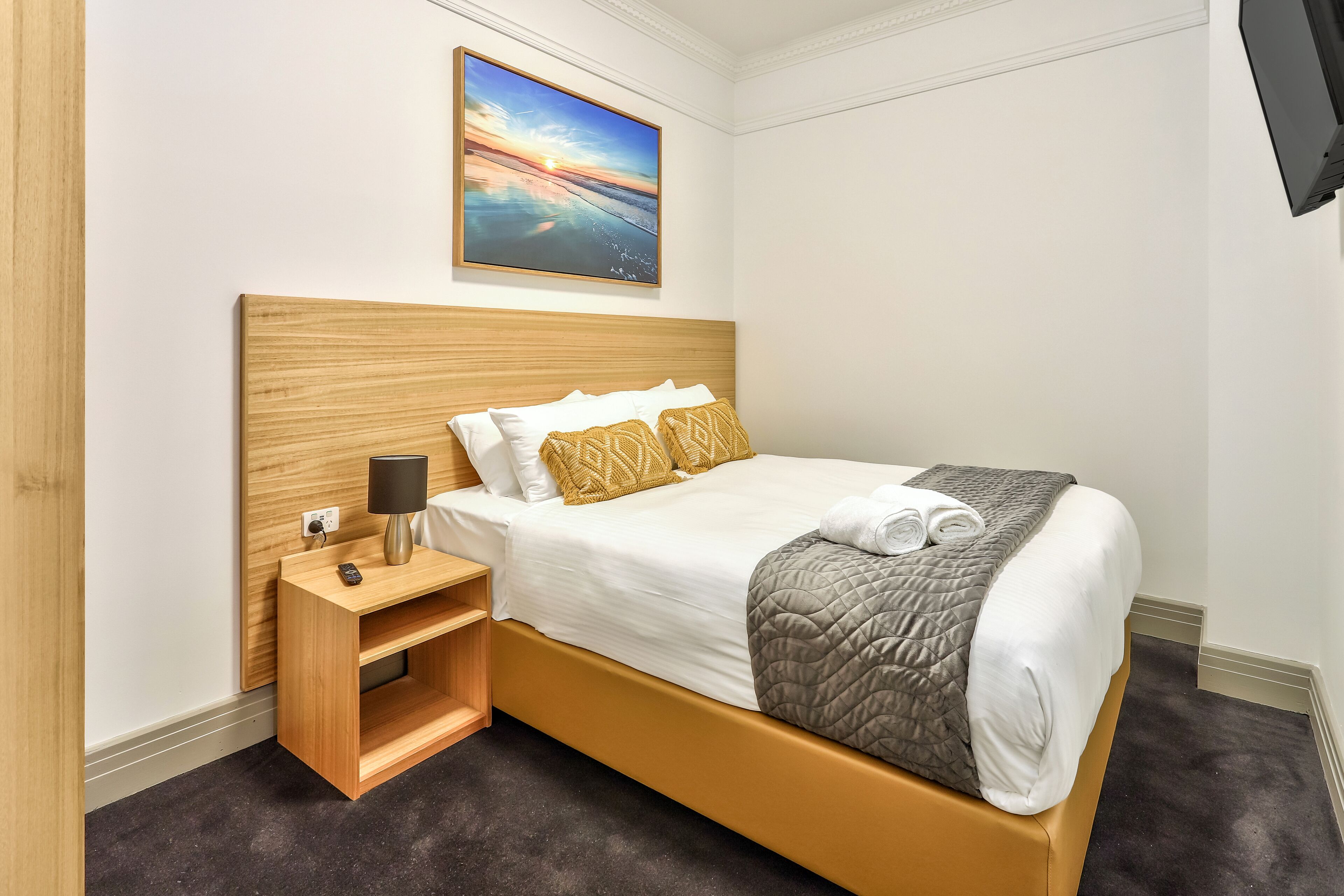 A hotel room with a wooden bed frame, white bedding, a nightstand with a lamp, and a painting of a sunset over the ocean on the wall.