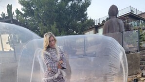 Une personne se tient devant une structure transparente en forme de dôme, dans un jardin où l’herbe est verte et où poussent des fleurs jaunes.