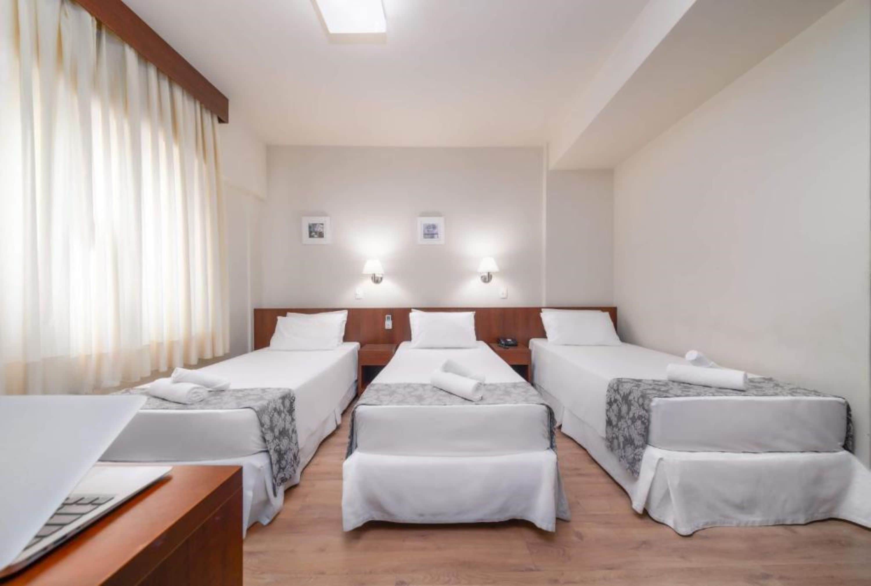 A hotel room with three single beds, a desk, and a large window with curtains.