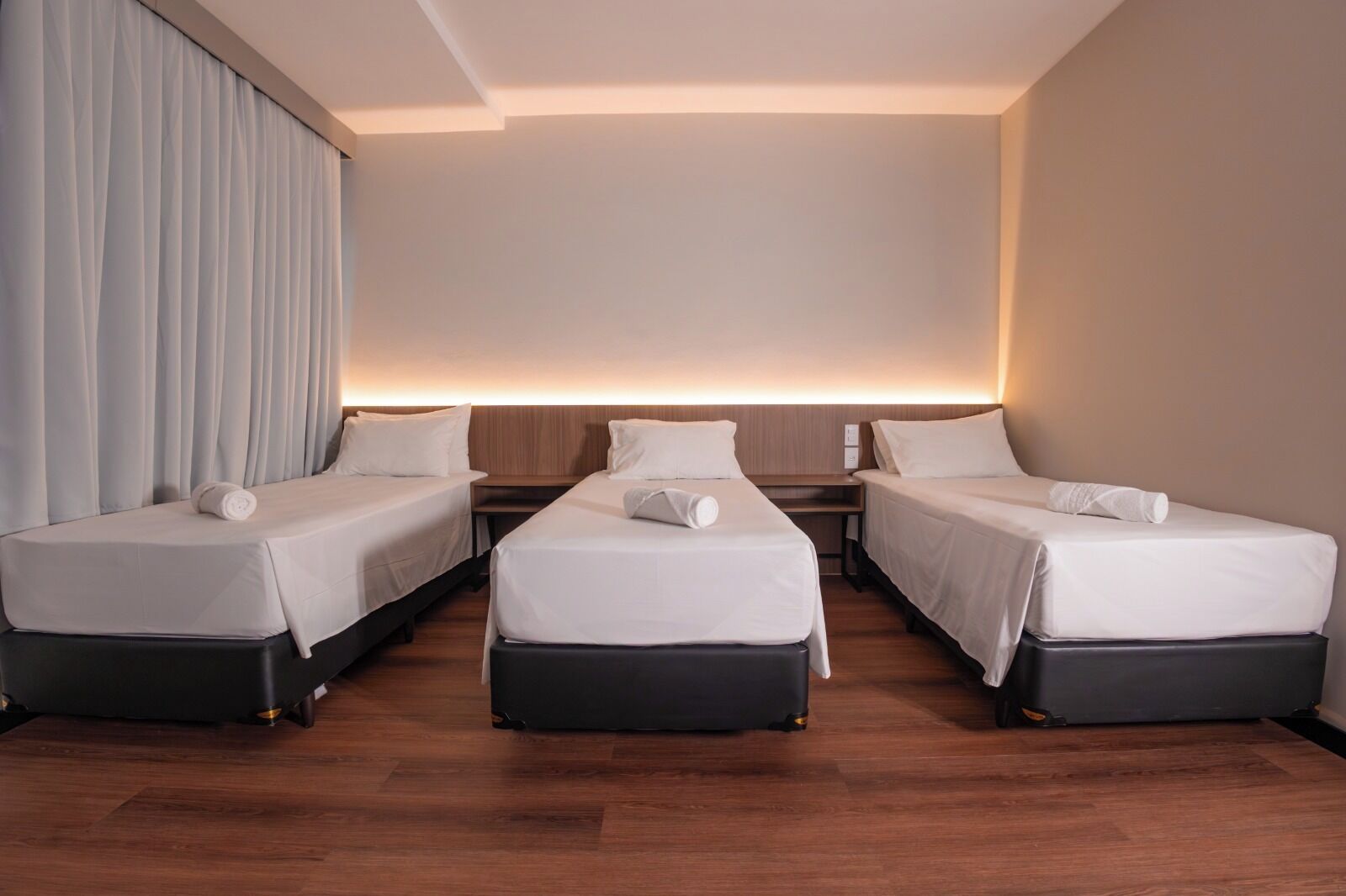 A hotel room with three single beds, a wooden floor, and a large window with curtains.