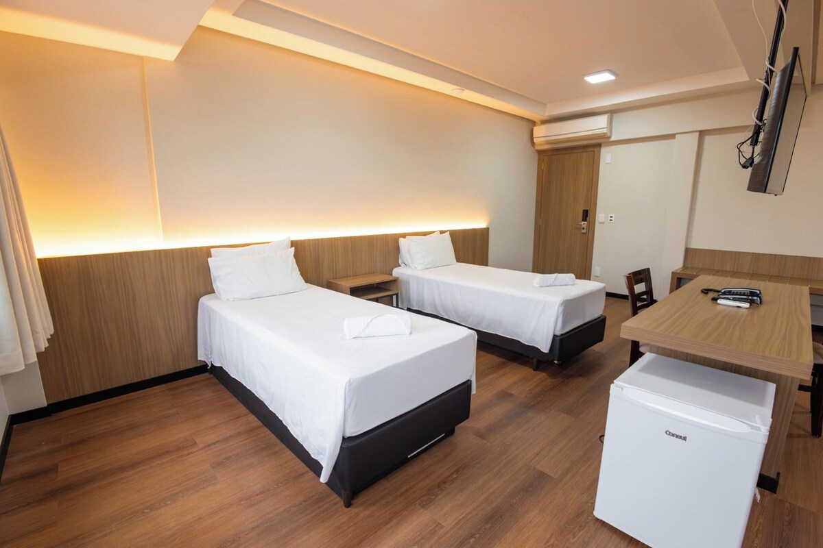 A hotel room with two beds, a desk, a chair, a small refrigerator, and a wall-mounted TV.