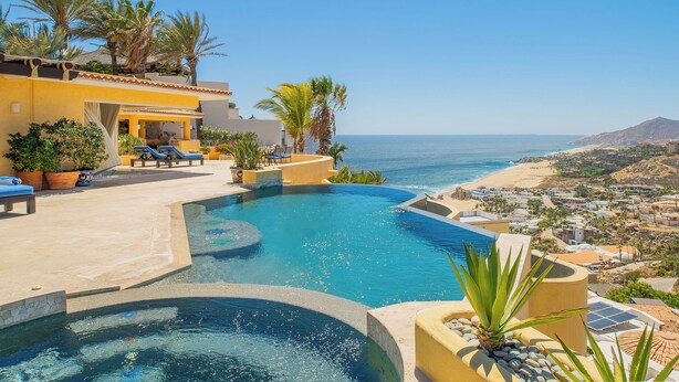 A pool with a view of the sea, palm trees, and a building with a yellow facade.