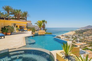 A pool with a view of the sea, palm trees, and a building with a yellow facade.