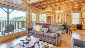A wooden cabin interior with a sofa, dining table, chairs, and a view through large windows. - 'Mountain Dreams' Cabin: Hot Tub, Sauna & Views! (Sevierville)