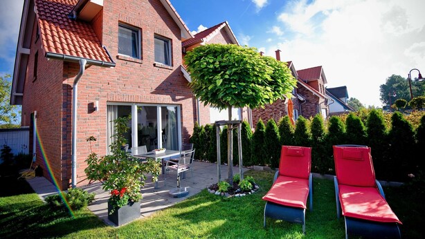 A two-story brick house with a red-tiled roof, a patio with a table and chairs, and a small garden with a tree and plants.