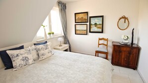 A bedroom with a large bed, a nightstand, a wooden chair, a dresser, and a window with curtains. There are three framed pictures on the wall.