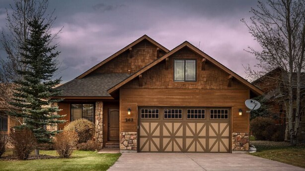 Une maison en bois à deux étages, avec des fondations en pierre, un garage doté d’une porte à motifs en losange et une antenne parabolique sur le toit.