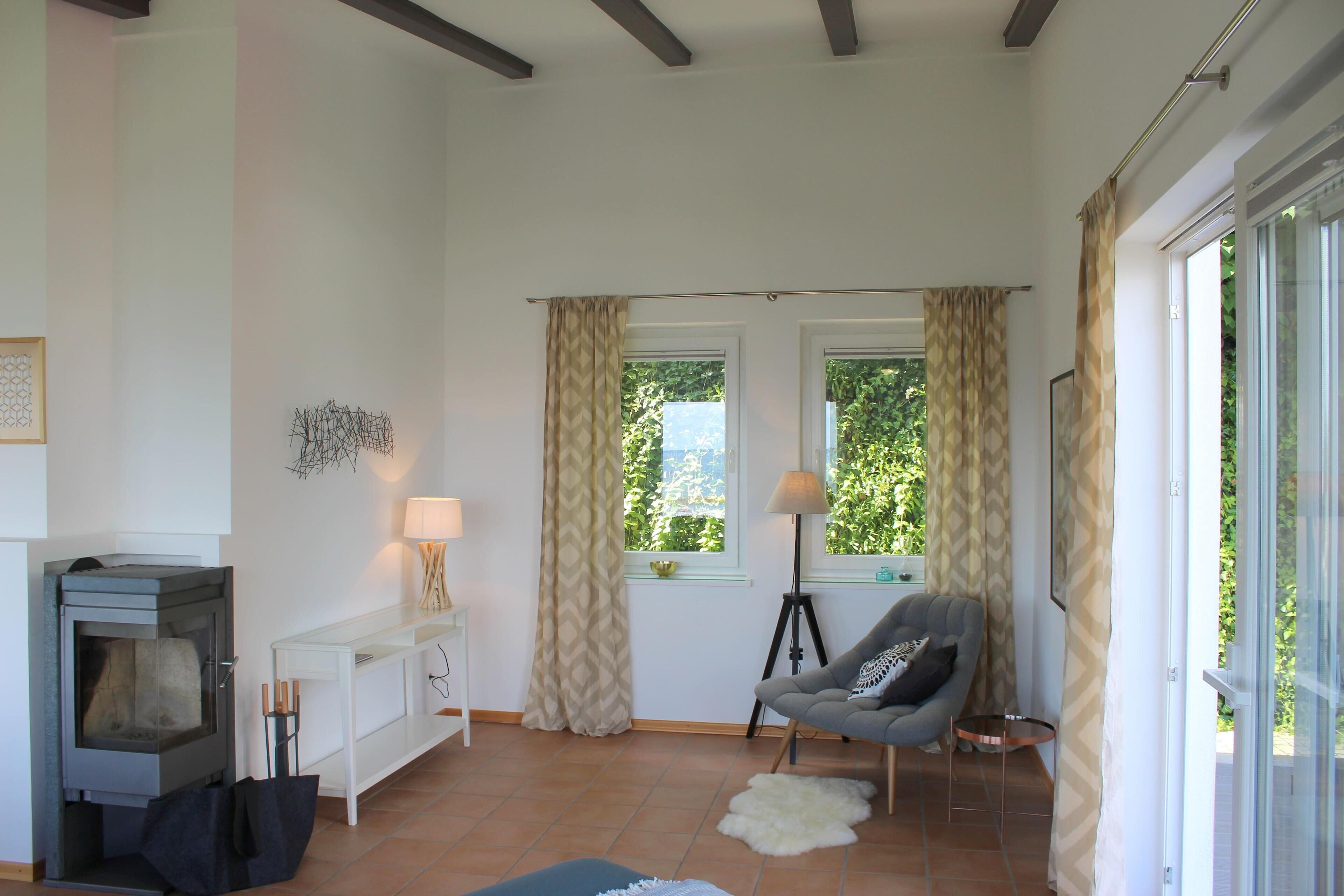 Ein Wohnzimmer mit Kamin, einer Couch, einer Lampe und Blick auf Grün hinter den Fenstern.