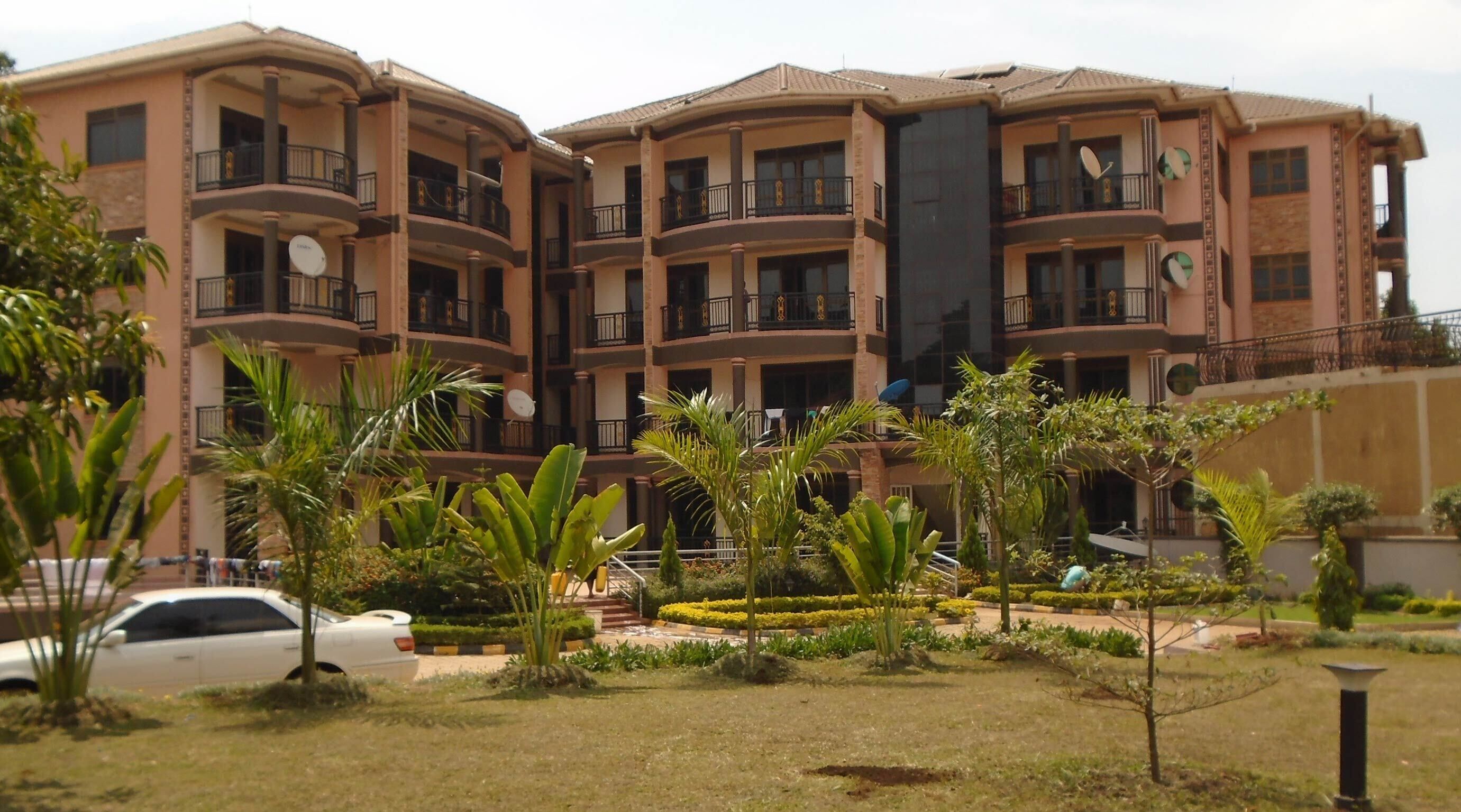 Un edificio residencial de varios pisos con balcones, rodeado de jardines cuidados y un automóvil estacionado.