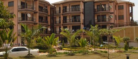 Un edificio residencial de varios pisos con balcones, rodeado de jardines cuidados y un automóvil estacionado.