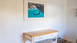 Une pièce avec un mur blanc, un tableau encadré représentant un paysage côtier, une étagère en bois et une armoire blanche avec une grille métallique en dessous.