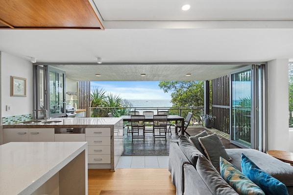 Una cocina moderna con vista al océano, un comedor con asientos exteriores y una sala de estar con sofá y cojines.